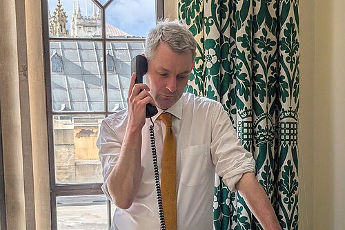 Will Forster speaking on the phone in his Westminster office, he is wearing a white shirt with the sleeves rolled up and a yellow tie.