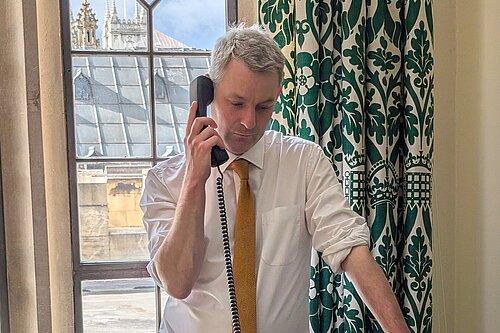 Will Forster speaking on the phone in his Westminster office, he is wearing a white shirt with the sleeves rolled up and a yellow tie.