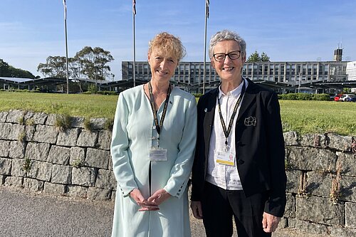 Cllrs Hillary Frank and Sarah Preece outside, with a stone wall, a grassy field with flagpoles and a building behind them