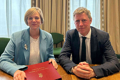 James seated at a table in front of some rather imposing curtains with Roads Minister Lilian Greenwood. She has an embossed folder in front of her.