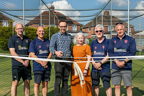 Steve Redwood, Scott Ewens, Adam Dance MP, Trudy Dyke, Rob Fox (Chairman), and Richard Reeves (President of Yeovil Cricket Club) stand together in a cricket training net area on a sunny day. Adam is holding scissors after cutting a ceremonial ribbon to mark the opening of a new cricket facility, with everyone smiling towards the camera.
