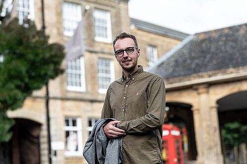 Adam Dance MP stands outside in a town centre, smiling at the camera. He is wearing a green button-up shirt and holding a grey coat. Behind him is a historic stone building and a traditional red phone box. The scene is bright with natural daylight.