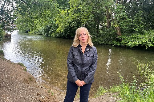 Liz Jarvis MP standing beside a river, wearing a black jacket and wellington boots. She is surrounded by trees and greenery, with a serious expression, highlighting concern for the local environment.