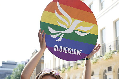 Someone holds up a round placard in Pride flag colours with a superimposed white Lib Dem bird-of-liberty and the hashtag #LoveIsLove