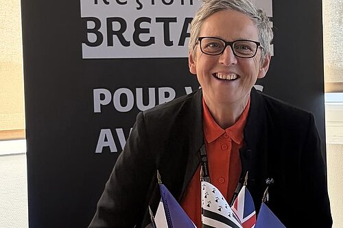 Cllr Sarah Preece in front of a banner saying 'Région Bretagne'. The tops of flags representing the Celtic nations, including Cornwall and Brittany, are just visible at the bottom