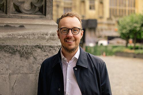 Adam Dance MP smiling in front of a stone pillar outside the Houses of Parliament, wearing glasses, a white shirt, and a dark jacket.