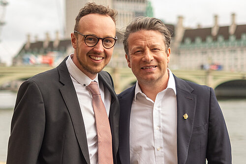 Adam Dance MP and Jamie Oliver stand side by side, smiling, with the River Thames and Westminster buildings in the background. Both are dressed in suits, with Adam wearing a pink tie and Jamie in an open-collared white shirt and dark blazer. The London Eye is partially visible on the left under a cloudy sky.