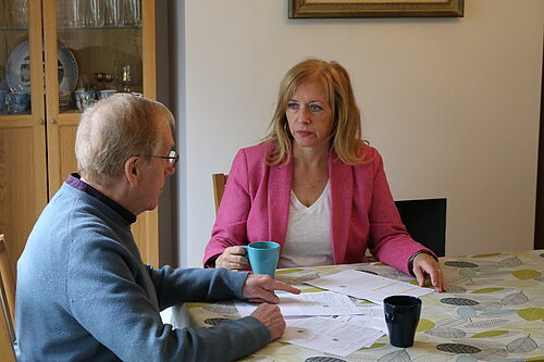 Liz Jarvis MP sitting at a kitchen table with an older man, discussing paperwork. She is wearing a pink blazer and holding a turquoise mug, while he points to documents spread out on the table. Both appear focused and engaged in conversation.