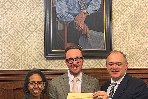 Adam Dance MP stands with two colleagues in Parliament holding a printed Bill, with a large painted portrait hanging on the wall behind them.