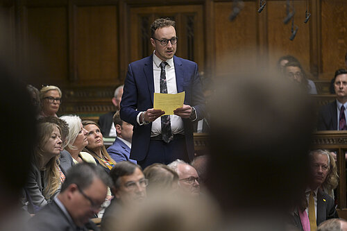 Adam Dance MP stands in the House of Commons, surrounded by MPs. He wears a blue suit and tie, a white shirt, and holds a small piece of paper.