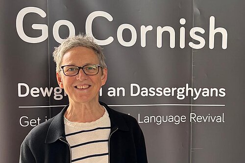 Cllr Sarah Preece in front of a banner saying 'Go Cornish', and two lines of partially obscured text; the first in Cornish: 'Dewg... an Dasserghyans', the second: 'Get... Language Revival'