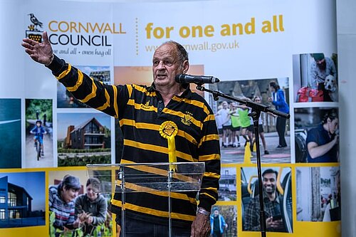 Cornwall Councillor Jim Candy, dressed in Cornish colours, gestures by a microphone. In the background is a collage of pictures, topped by Cornwall Council for one and all.