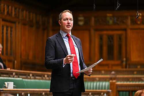Richard Foord standing up and speaking in the House of Commons. He is wearing a black suit with a red tie