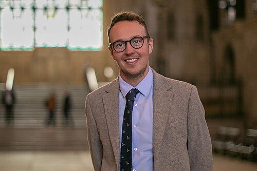 Adam Dance MP stands smiling inside Westminster Hall, part of the Houses of Parliament. He wears a light grey suit, blue shirt, and navy tie with a subtle pattern. Behind him, the grand arched window and stone staircase are softly out of focus, highlighting the historic setting.