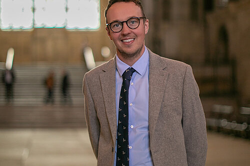Adam Dance MP stands smiling inside Westminster Hall, part of the Houses of Parliament. He wears a light grey suit, blue shirt, and navy tie with a subtle pattern. Behind him, the grand arched window and stone staircase are softly out of focus, highlighting the historic setting.