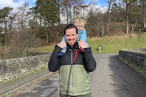 Adam Harley outside in a waterproof jacked with his son on his shoulders holding his hands.