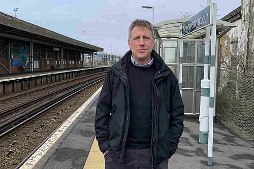 James MacCleary standing on the platform at Newhaven Harbour station. Behind him are empty tracks, station buildings and signalling paraphrenalia