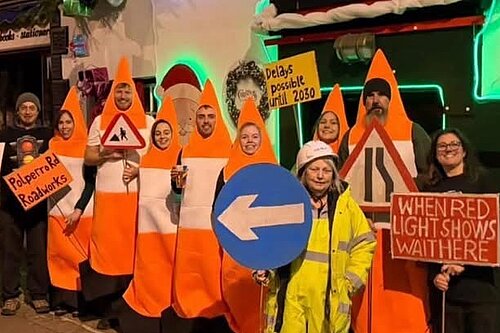 People dressed as traffic cones stand outside a pub. They are holding a variety of placards designed to resemble road signs.