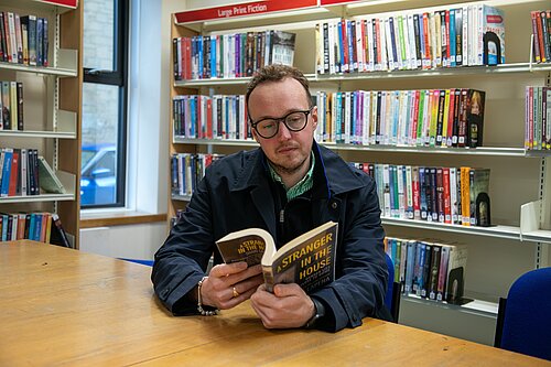 A man sits at a table in a library reading a book, with shelves of books behind him.