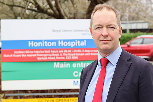 A photo of Richard Foord wearing a suit and standing in front of the sign for Honiton Hospital, which is out of focus