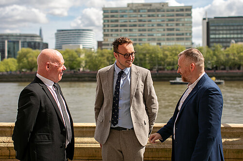 Peter Brown (Technical & Development Director at the CPA), Adam Dance MP, and Steve Mulholland (CPA Chief Executive) stand together outside by the River Thames, engaged in conversation, with tall buildings and trees in the background.
