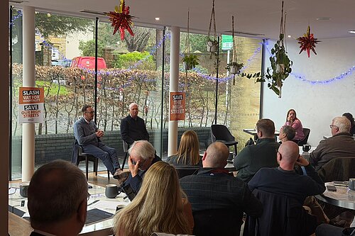 A group of people sit in a café style room listening to two men speaking at the front. Posters on the windows read Slash VAT for Hospitality and Save Our High Streets. Christmas decorations and hanging plants are visible.